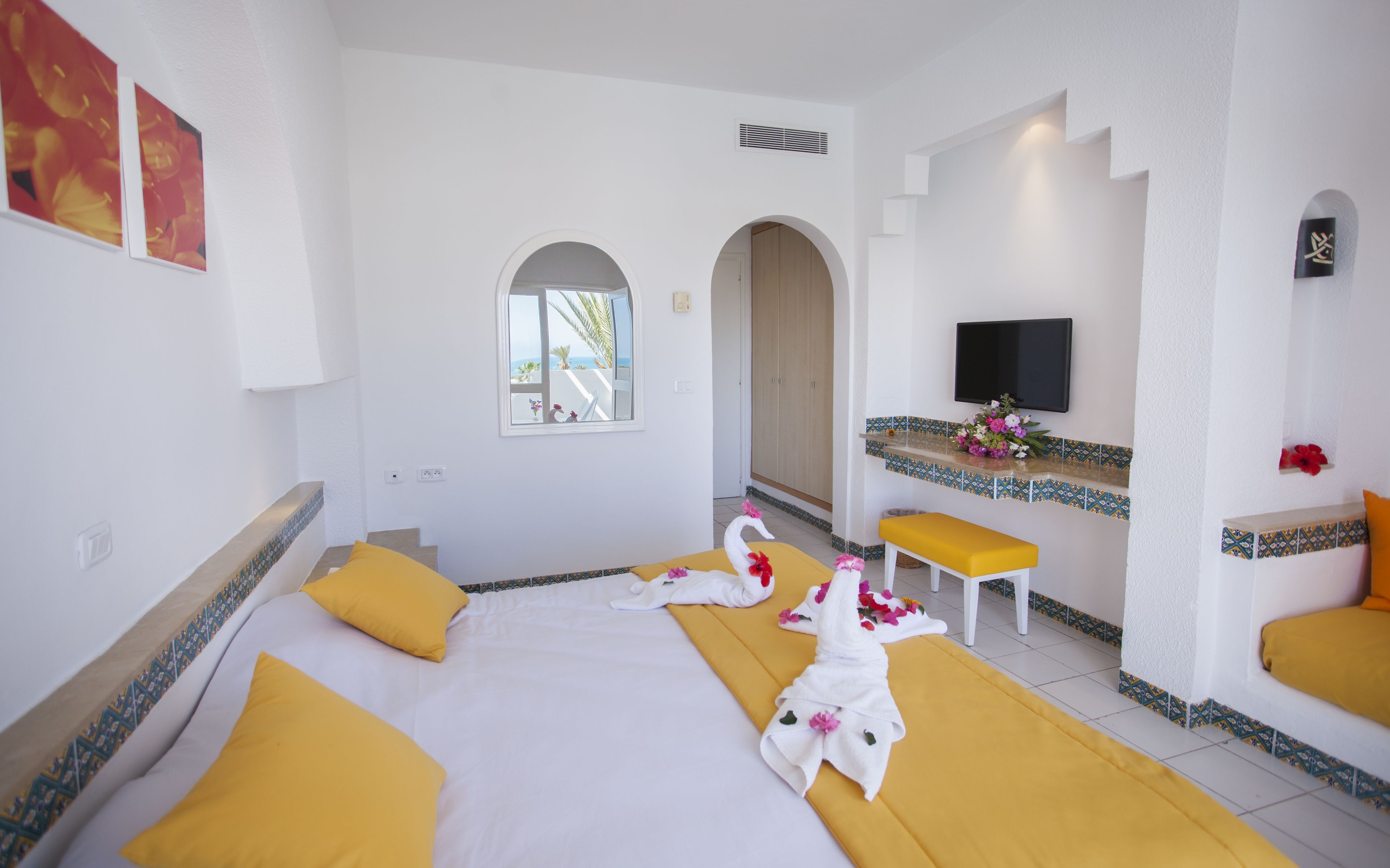 A hotel room with a large bed, yellow pillows, a small bench, a TV, and a window with a view.