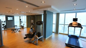 A modern gym with exercise equipment, large windows, and a view of the city. - AlHamra Hotel (Sharjah)