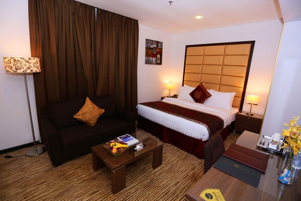 A hotel room with a bed, a sofa, a coffee table, and a desk with a lamp and flowers.