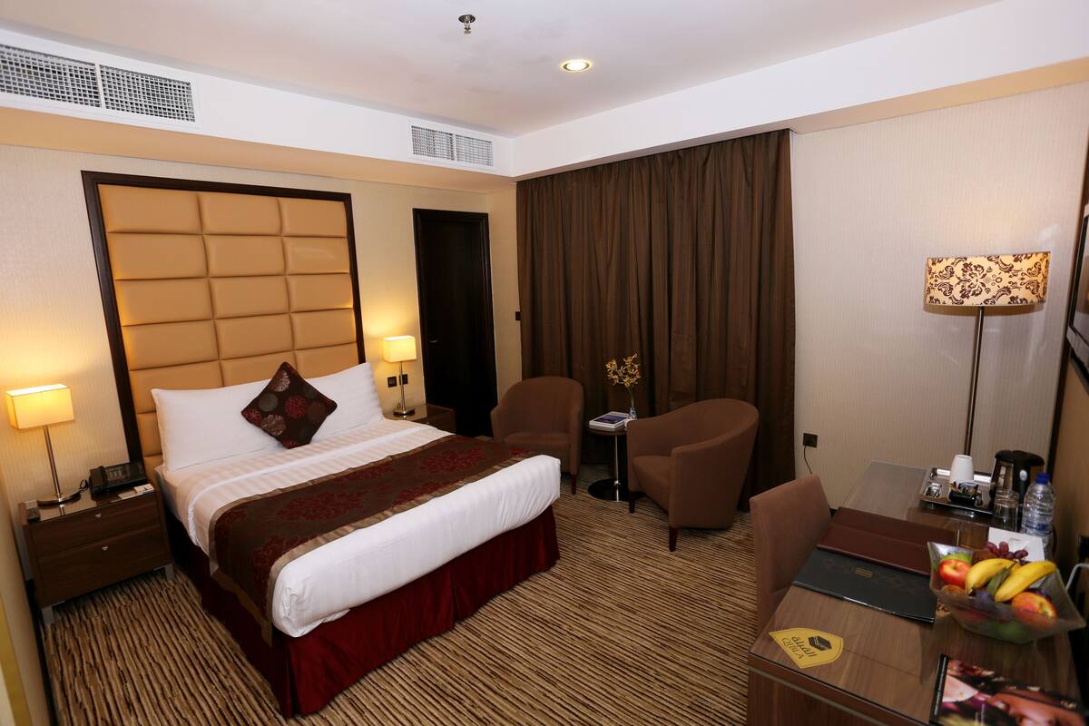 A hotel room with a bed, two chairs, a desk with a laptop, a fruit bowl, and a lamp.