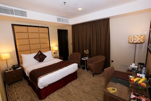 Une chambre d’hôtel avec un lit, deux chaises, un bureau avec un ordinateur portable, un panier de fruits et une lampe.