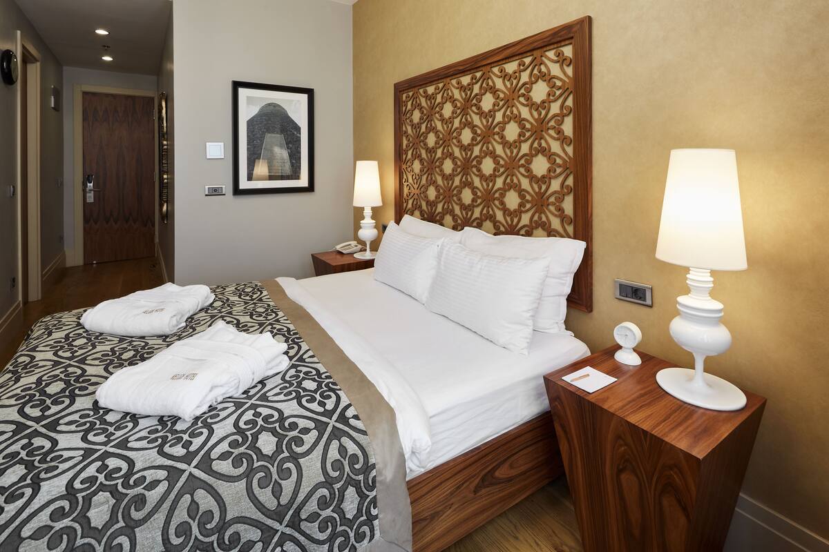 A hotel room with a large bed, two bedside tables with lamps, a wooden headboard, a framed picture on the wall, and a door in the background.