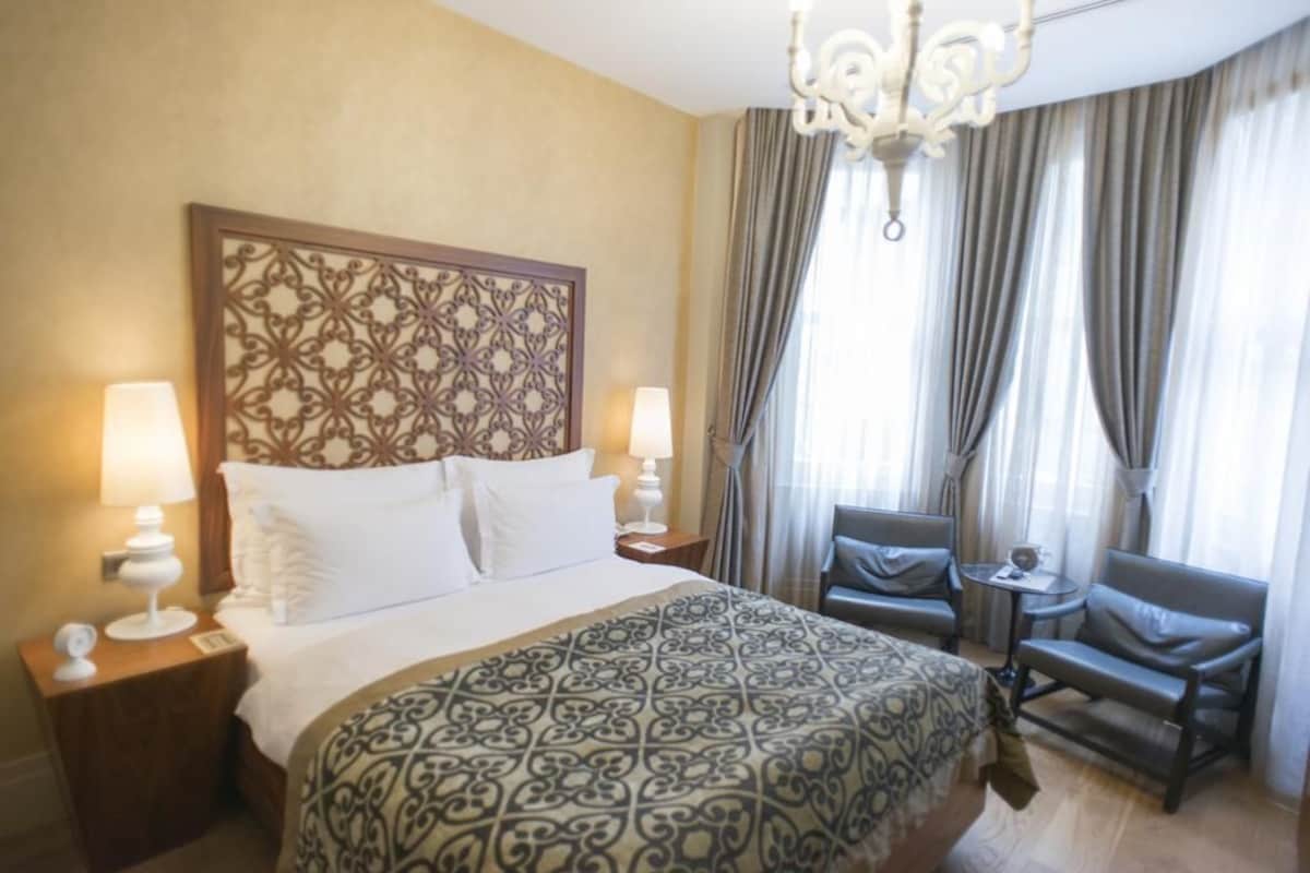 A hotel room with a large bed, two bedside lamps, a chandelier, a patterned bedspread, a decorative headboard, a seating area with two chairs, and a window with curtains.