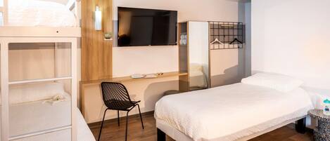 A hotel room with bunk beds, a desk with a computer, a chair, a bed, a TV, and a small table with a cup.