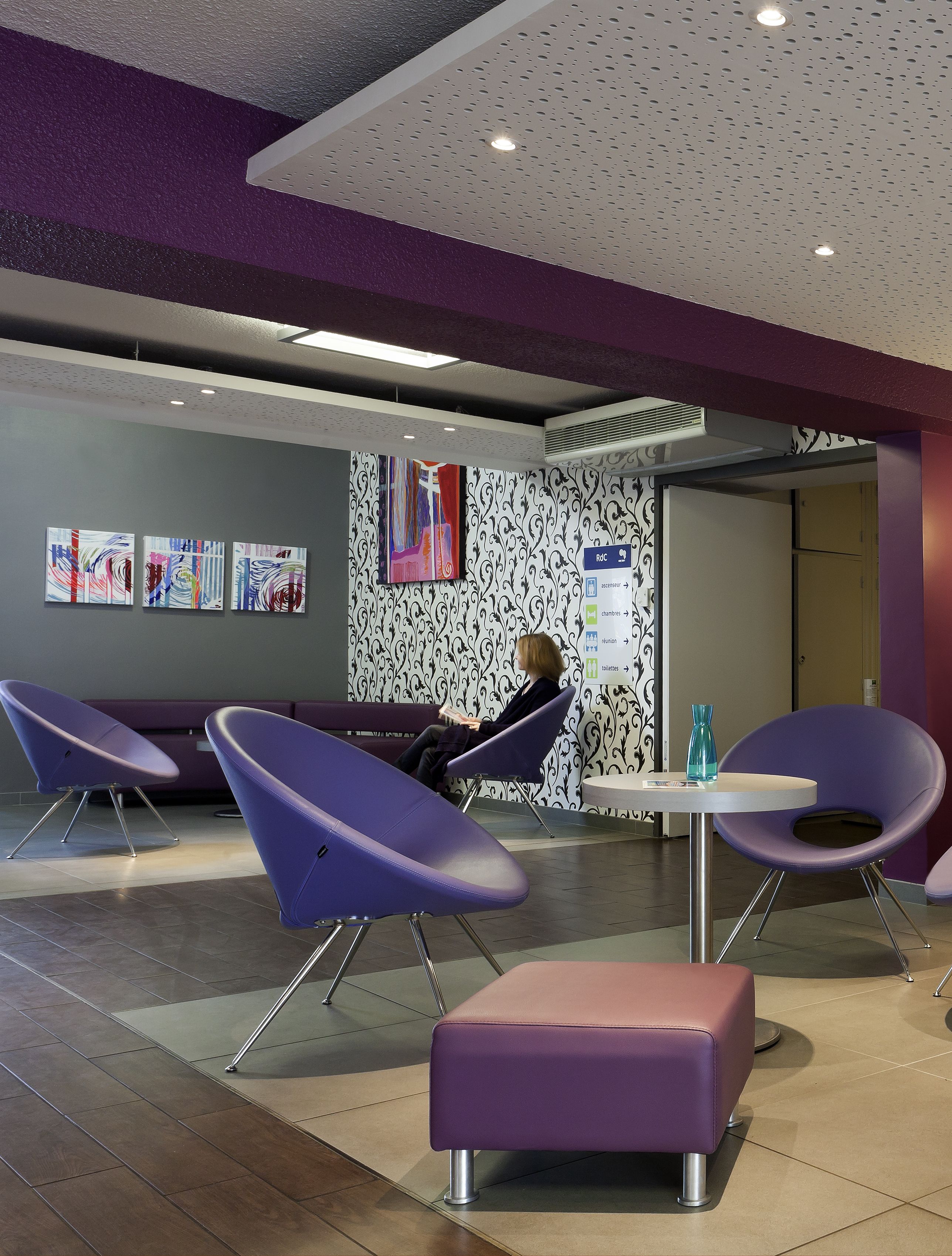 Un área de espera moderna con sillas moradas, un banco y una mesa con un florero. Se observan obras de arte en las paredes y un papel tapiz con diseño detrás de los asientos.