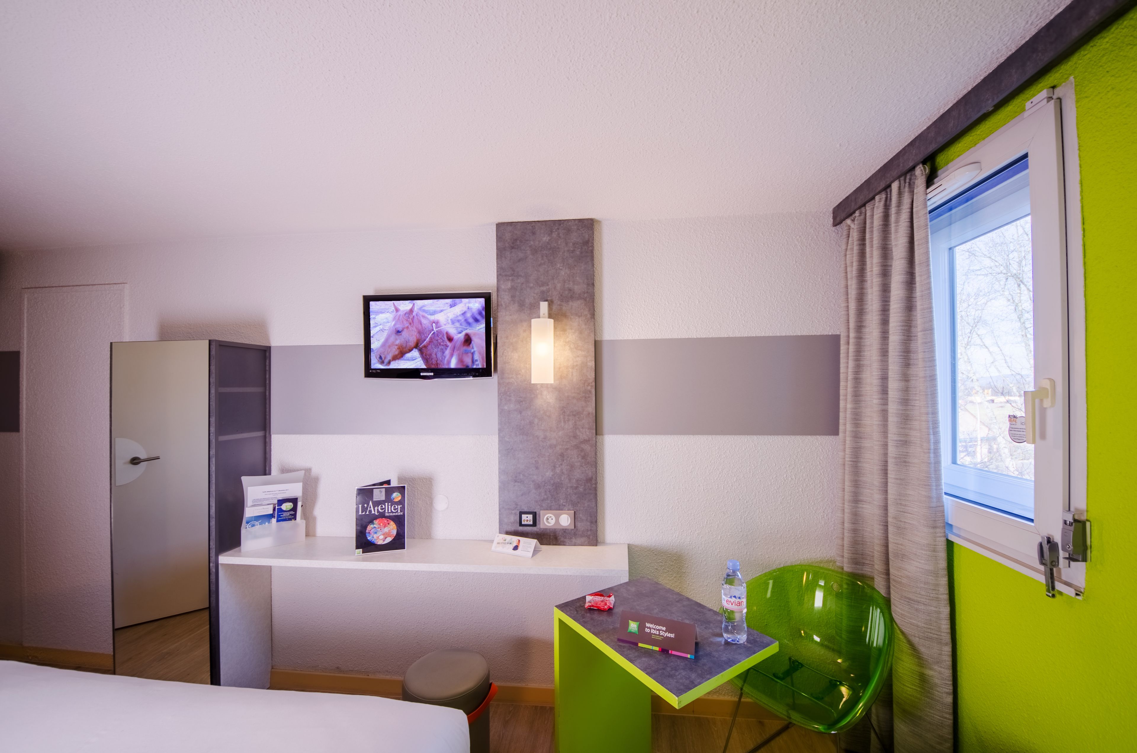 Une chambre d’hôtel avec un lit, un bureau avec un ordinateur, une télévision, un fauteuil vert et une fenêtre avec des rideaux.