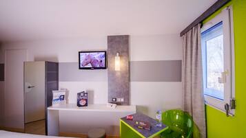 Une chambre d’hôtel avec un lit, un bureau avec un ordinateur, une télévision, un fauteuil vert et une fenêtre avec des rideaux.