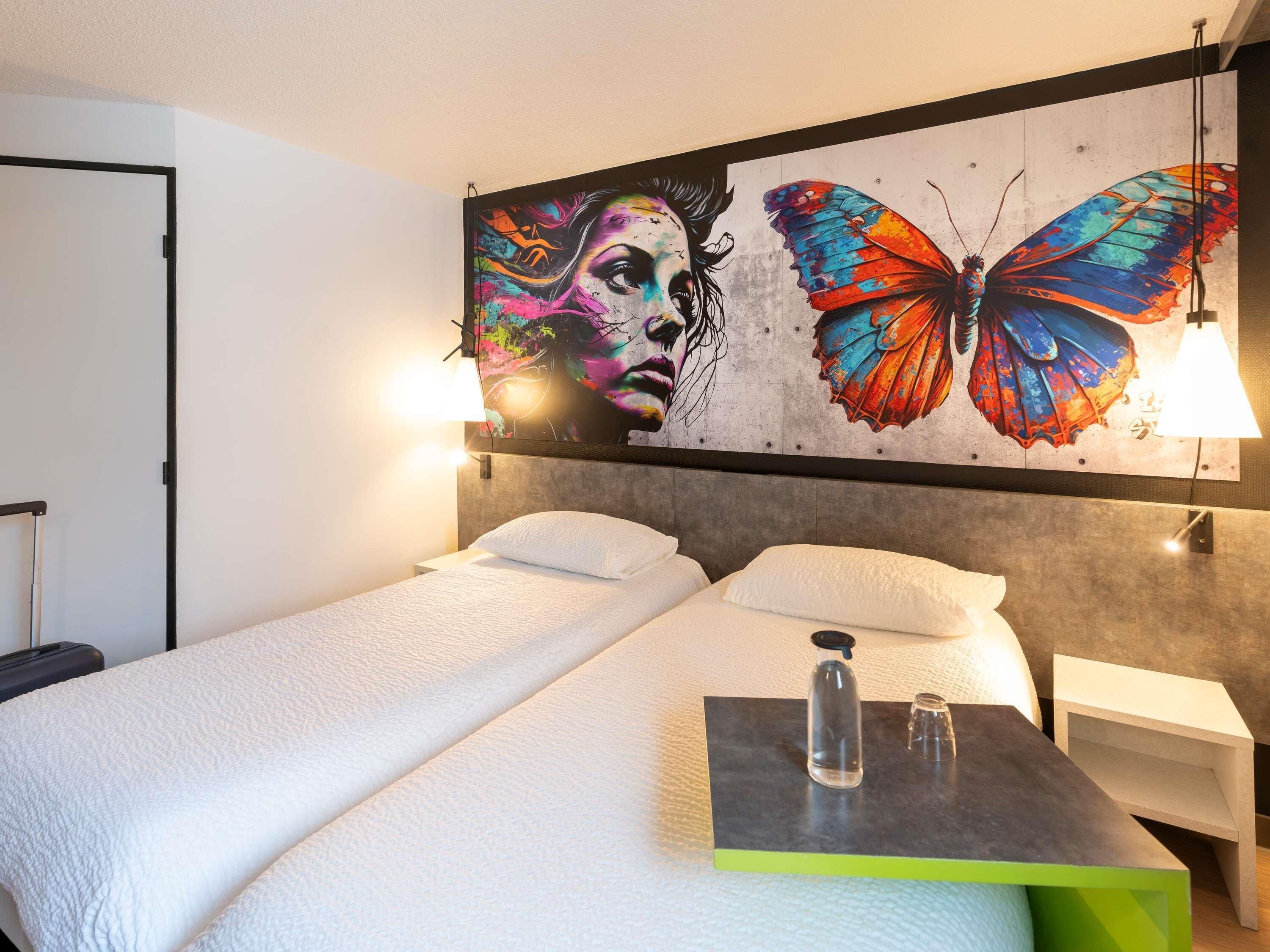Habitación de hotel con cama, mesita de noche, lámpara de pared y un mural de mariposas coloridas.