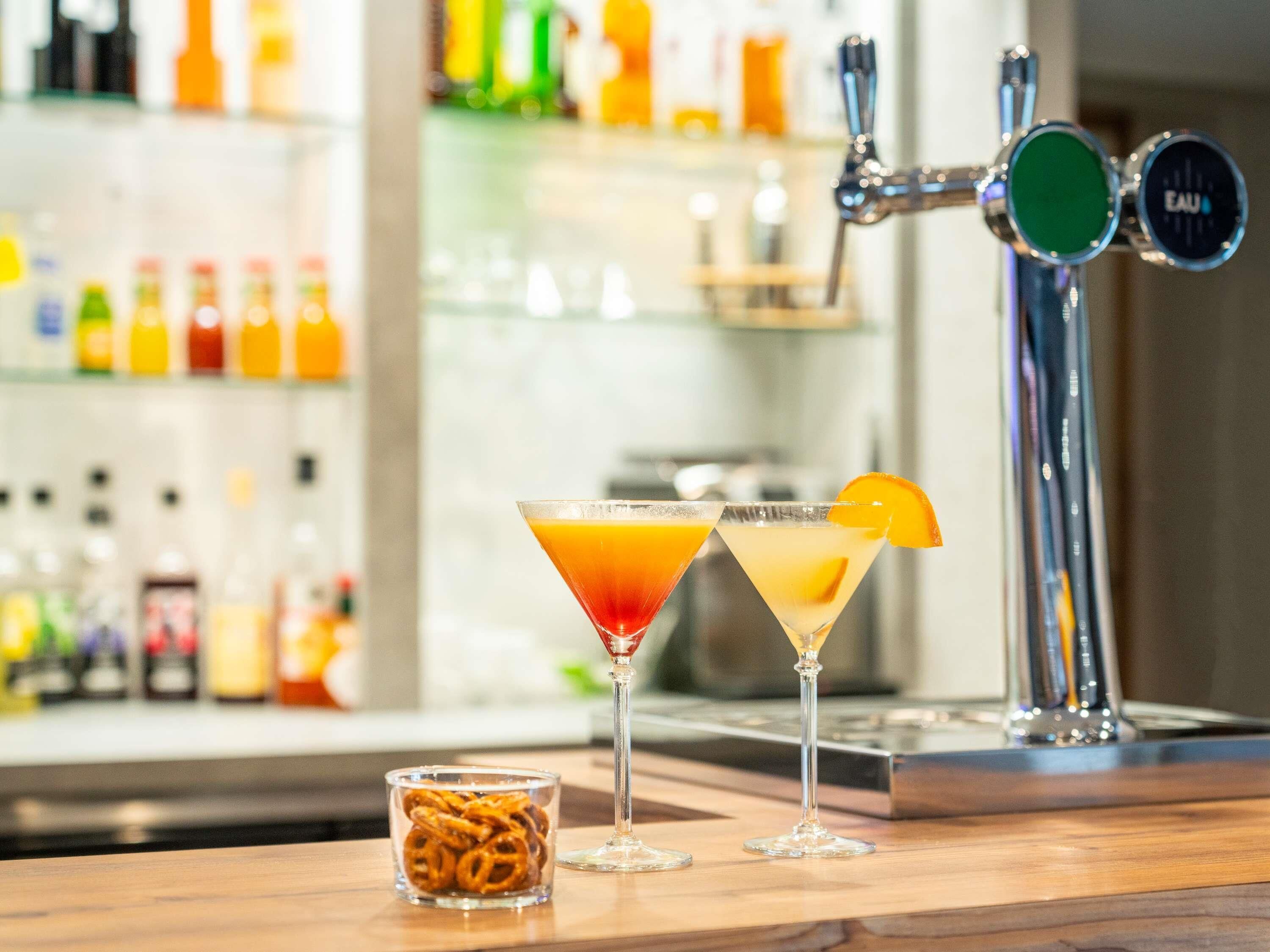 Two martini glasses with orange garnishes on a bar counter, a small bowl of pretzels, and a bar tap labeled "EAU".