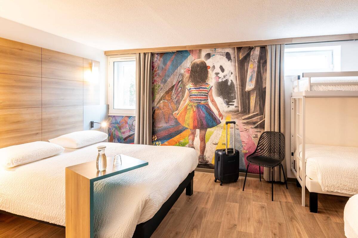 A hotel room with a bed, a desk, a chair, a suitcase, and a mural on the wall.