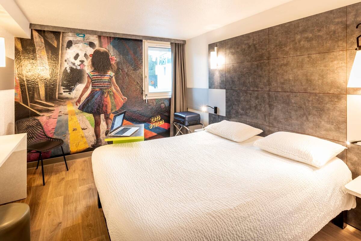 A hotel room with a large bed, a desk with a laptop, a chair, and a wall mural.