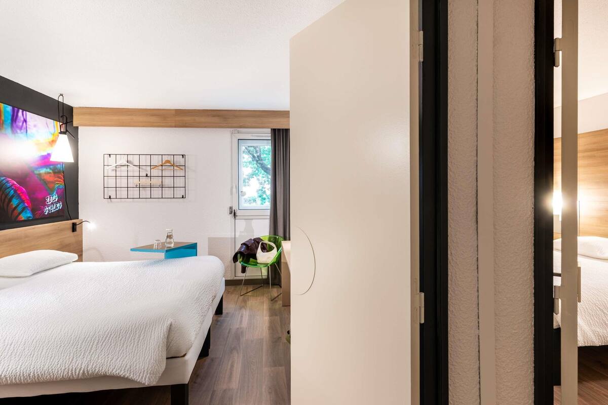 A modern hotel room with a large bed, a small desk, a chair, and a window with a view of trees.