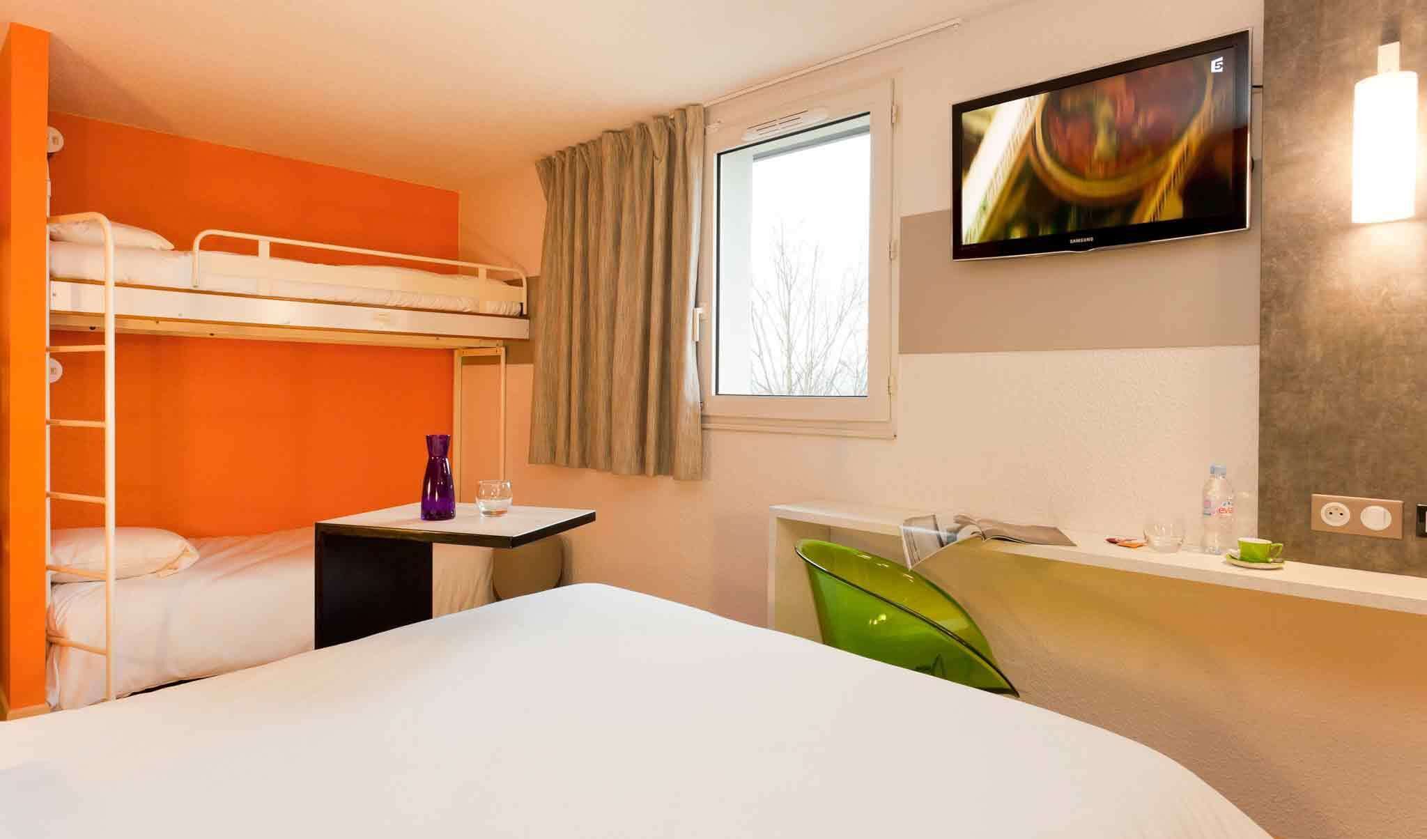 A hotel room with a bunk bed, a desk with a chair, a TV mounted on the wall, and a window with curtains.