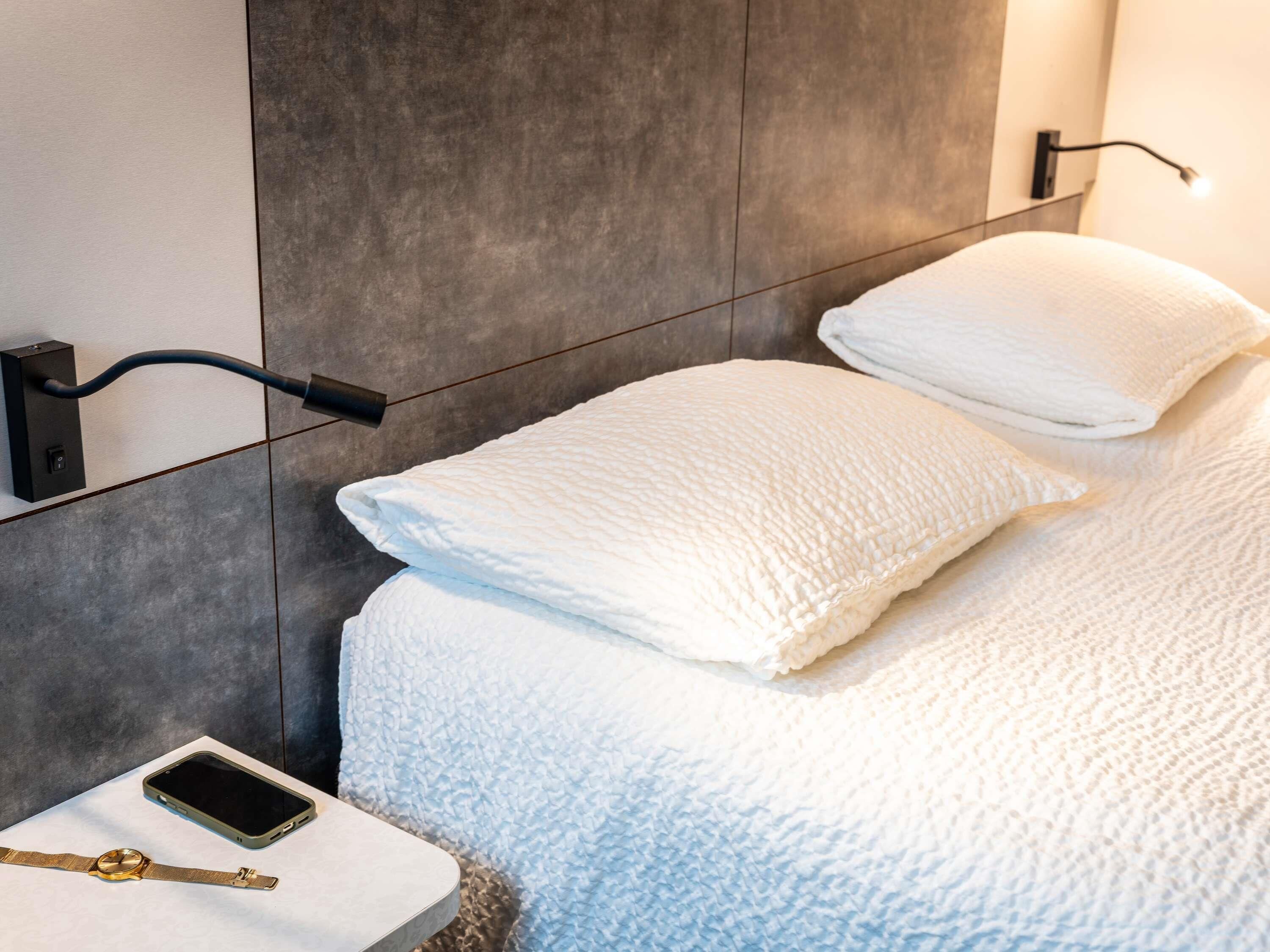A modern hotel room with a bed, white pillows, a bedside table with a smartphone and a watch, and a wall-mounted light fixture.