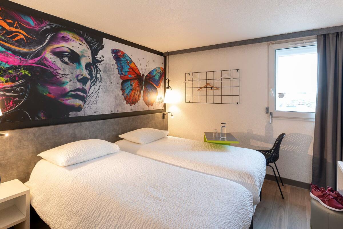 A hotel room with two beds, a large wall mural, a small table with a bottle and glass, and a chair.