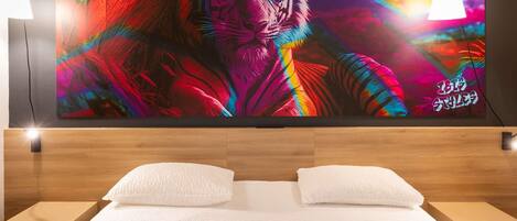 A bedroom with a large wall-mounted TV displaying a colorful tiger image, two bedside lamps, a neatly made bed with white bedding, and a small bedside table with a glass.
