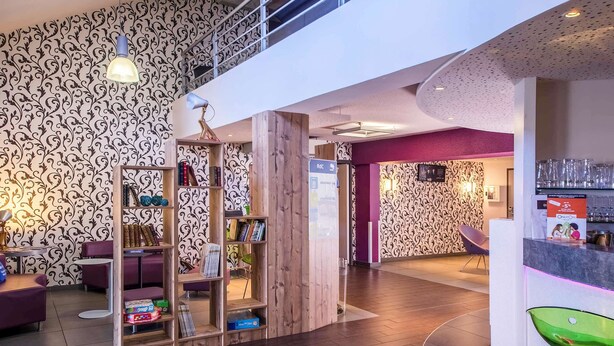 A modern lobby with a wooden bookshelf, purple seating, and a reception desk with a glass display. - ibis Styles Brive La Gaillarde (Brive-la-Gaillarde)