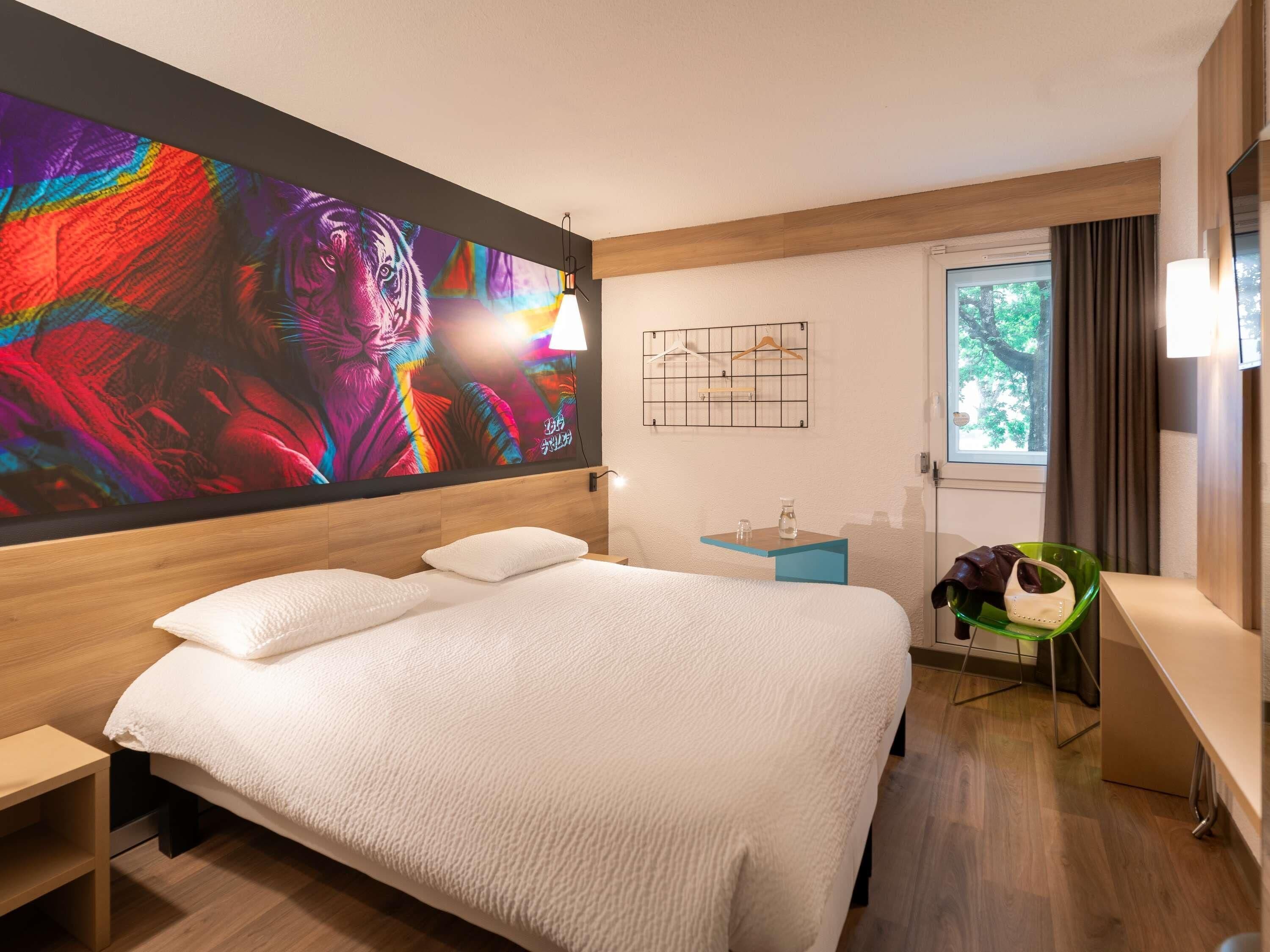 A modern hotel room with a large bed, a bedside table, a small side table, a green chair, and a window with curtains.