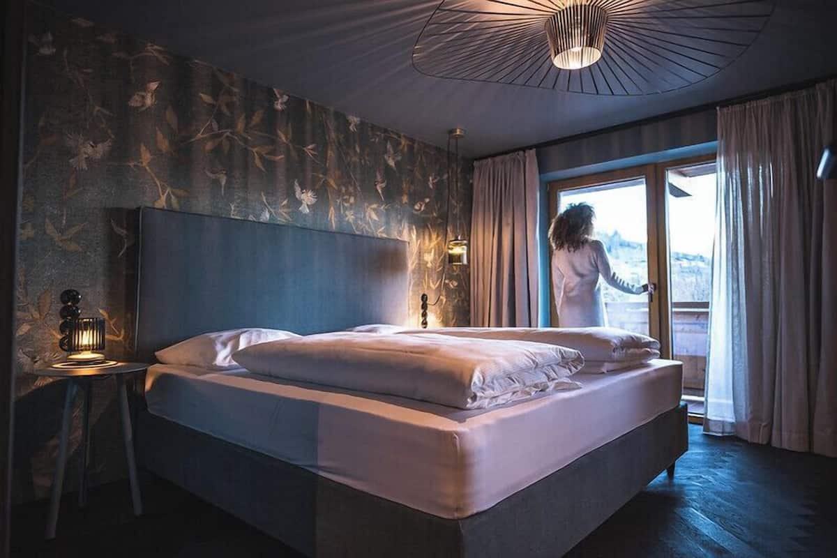 A modern hotel room with a large bed, a nightstand with a lamp, and a view of the outdoors through a sliding door.
