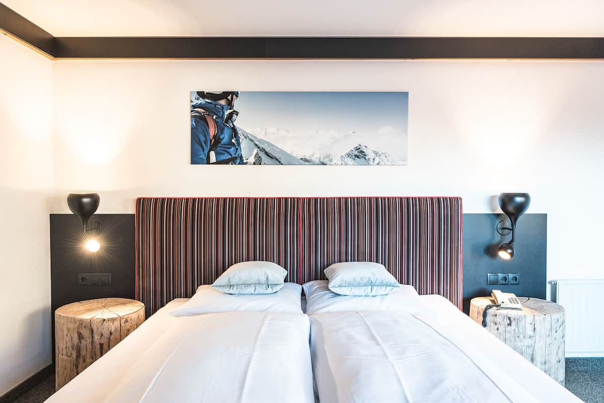 A hotel room with a large bed, two bedside tables, a headboard with vertical stripes, a painting of a mountain, and a wall-mounted lamp.