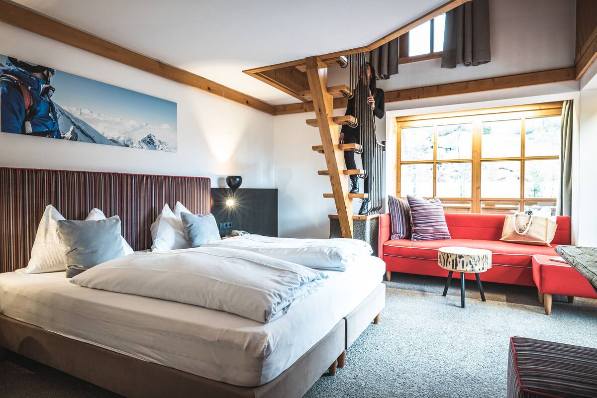 A bedroom with a large bed, a red sofa, a wooden ladder, and a window with a view.