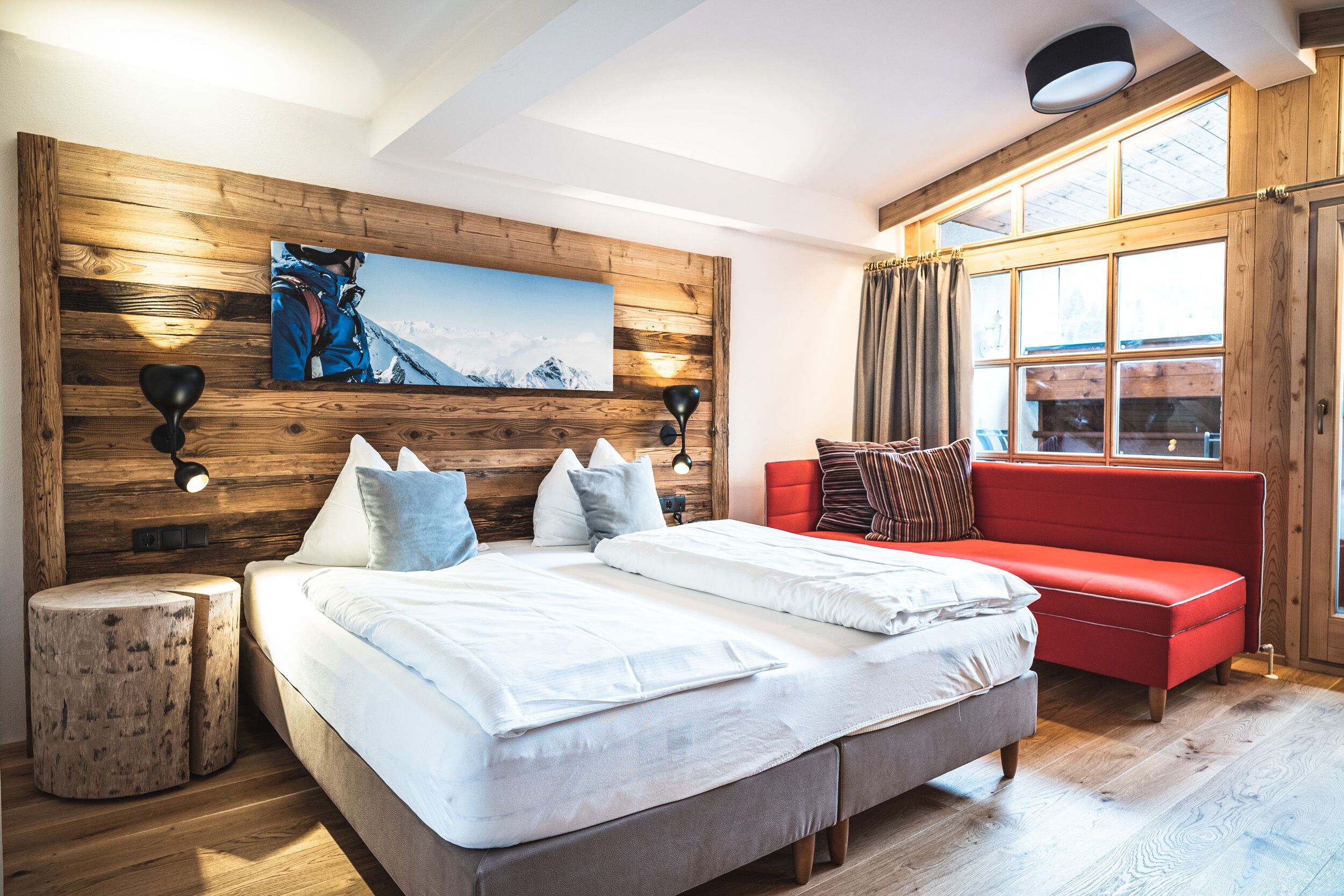 A modern bedroom with a wooden headboard, a large bed with white bedding, a red sofa, and a mountain painting on the wall.