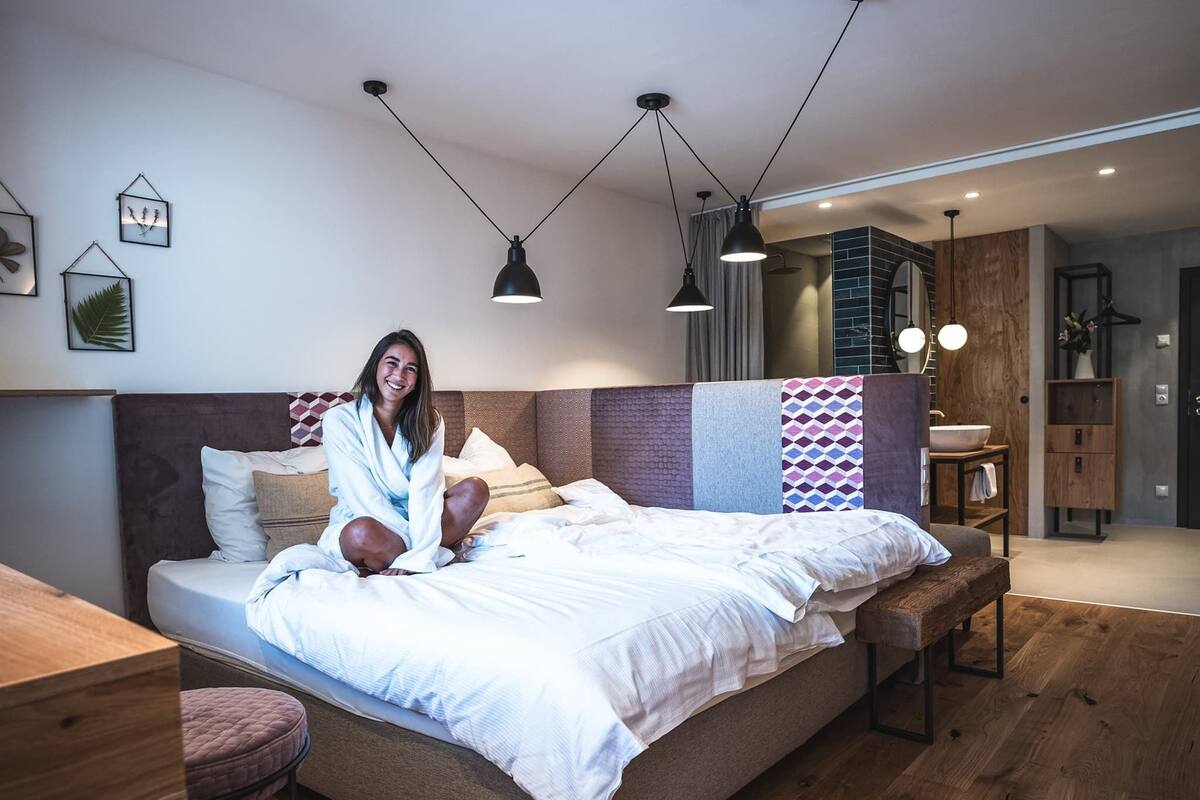 A modern bedroom with a large bed, a wooden bench, a bedside table, and a bathroom visible in the background.
