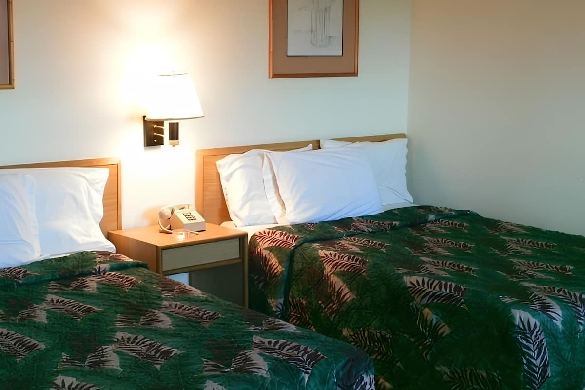 A hotel room with two beds, a nightstand with a phone, and a wall-mounted lamp.