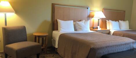 A hotel room with two beds, a chair, a small table, and a lamp.