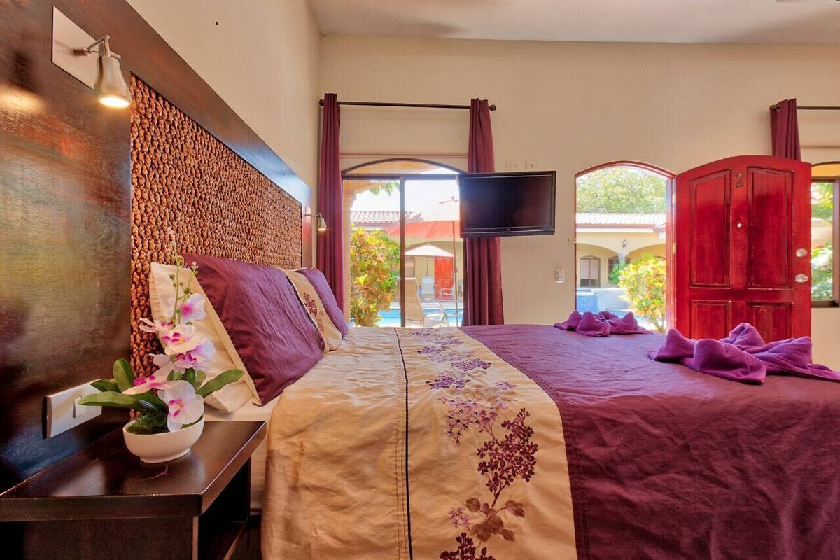 A hotel room with a large bed, a flat-screen TV mounted on the wall, and a view of the outside through a glass door.