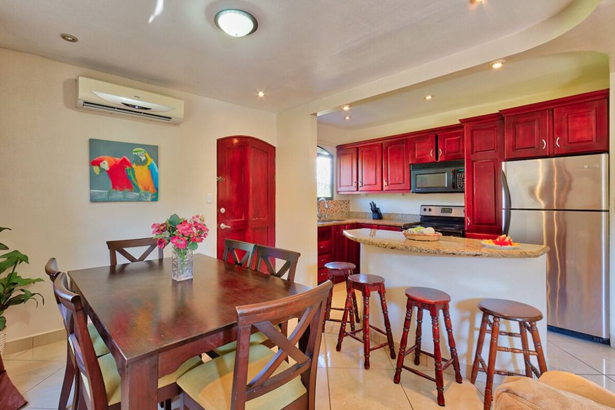 A kitchen with red cabinets, a stainless steel refrigerator, and a dining area with wooden chairs and a vase of flowers.