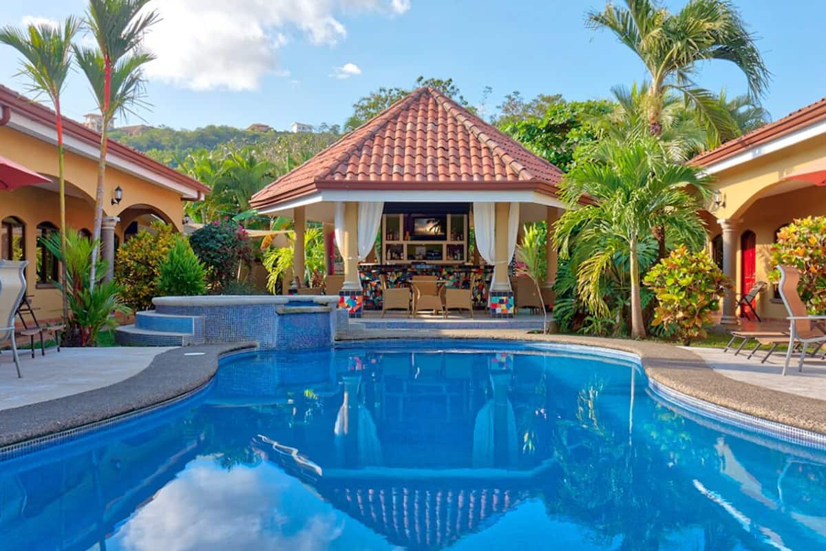 A swimming pool with a tiled deck, a gazebo with a tiled roof, and palm trees.