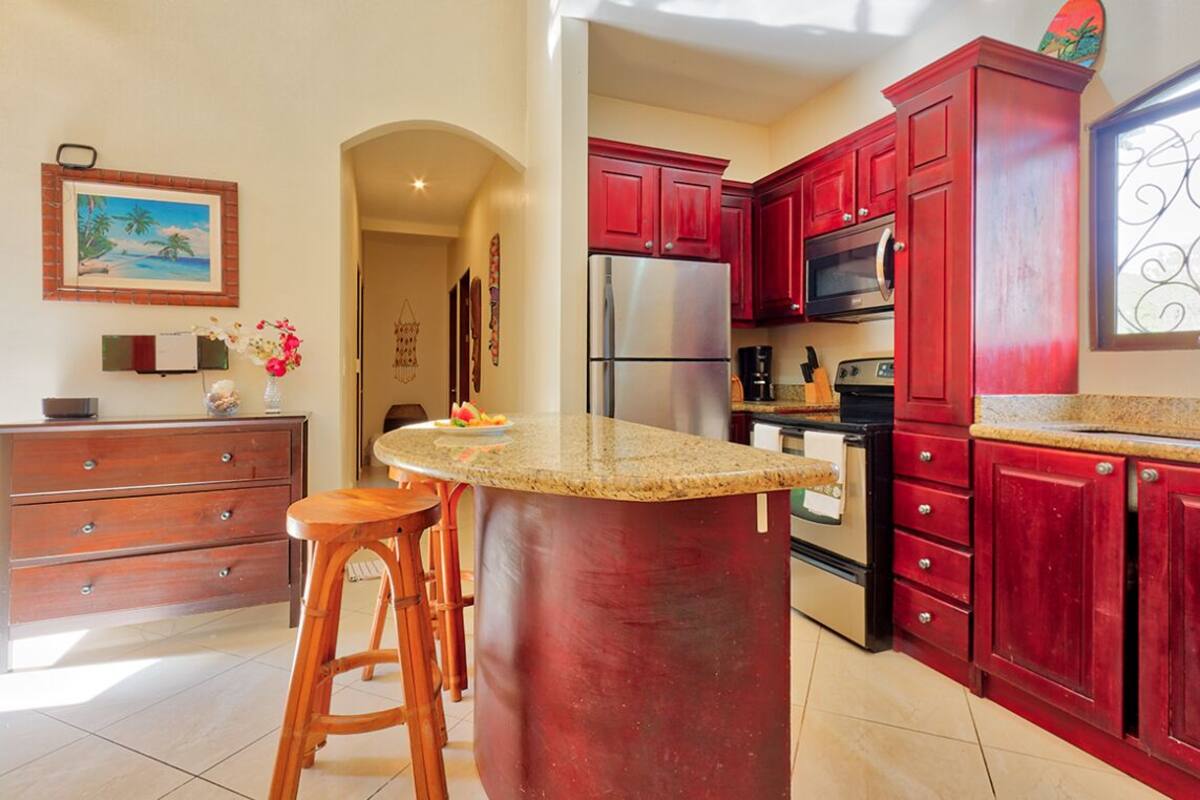 A kitchen with red cabinets, a stainless steel refrigerator, a microwave, and a coffee maker. There is a wooden stool and a small island with a granite countertop.