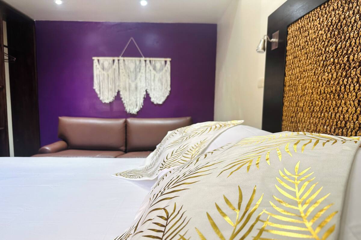 A bedroom with a purple wall, a hanging macrame piece, a brown leather sofa, and a bed with white and gold leaf-patterned pillows.