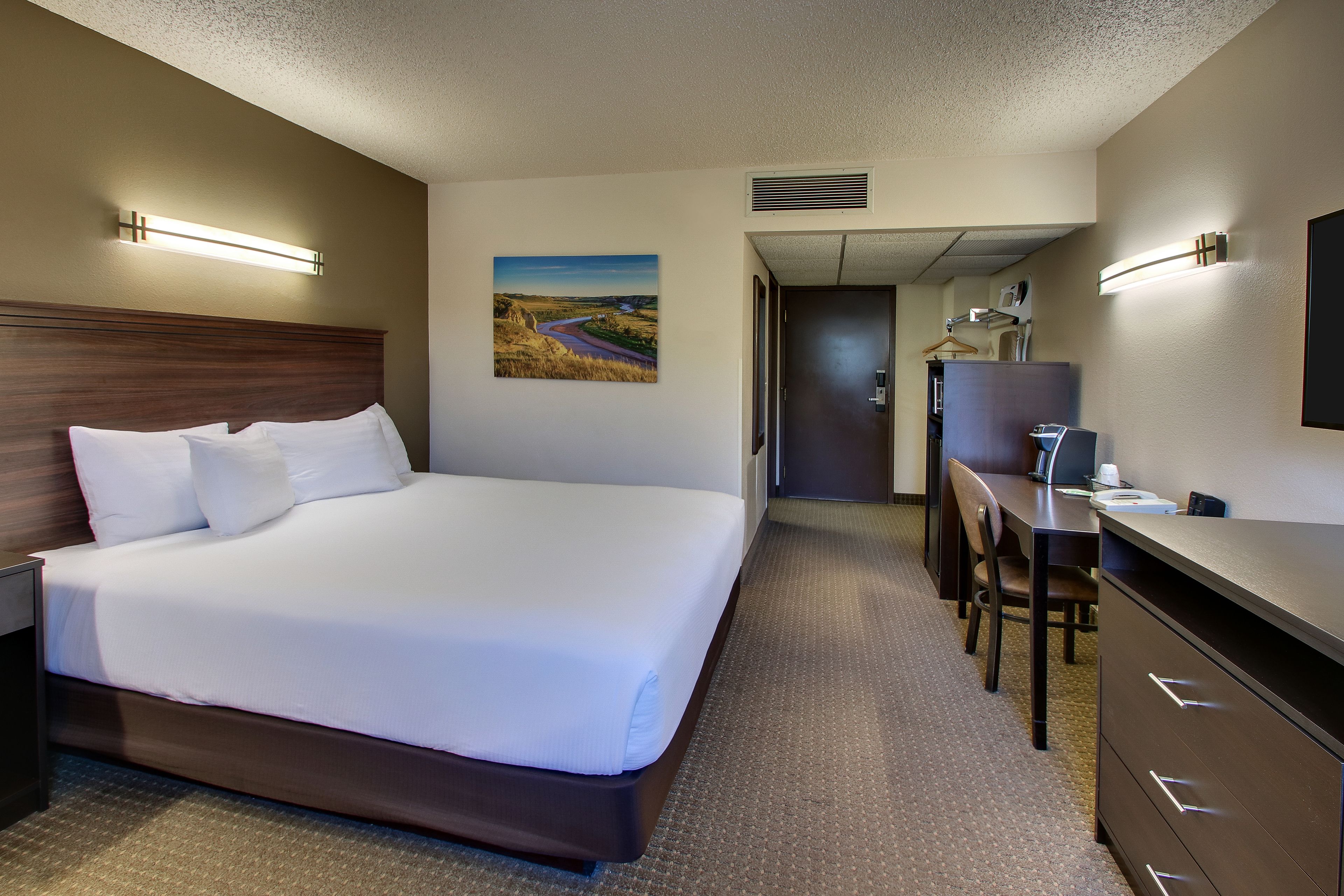 Ein Hotelzimmer mit einem großen Bett, einem Schreibtisch und einem Sessel. Ein Gemälde hängt an der Wand, und eine Tür führt in einen weiteren Raum.