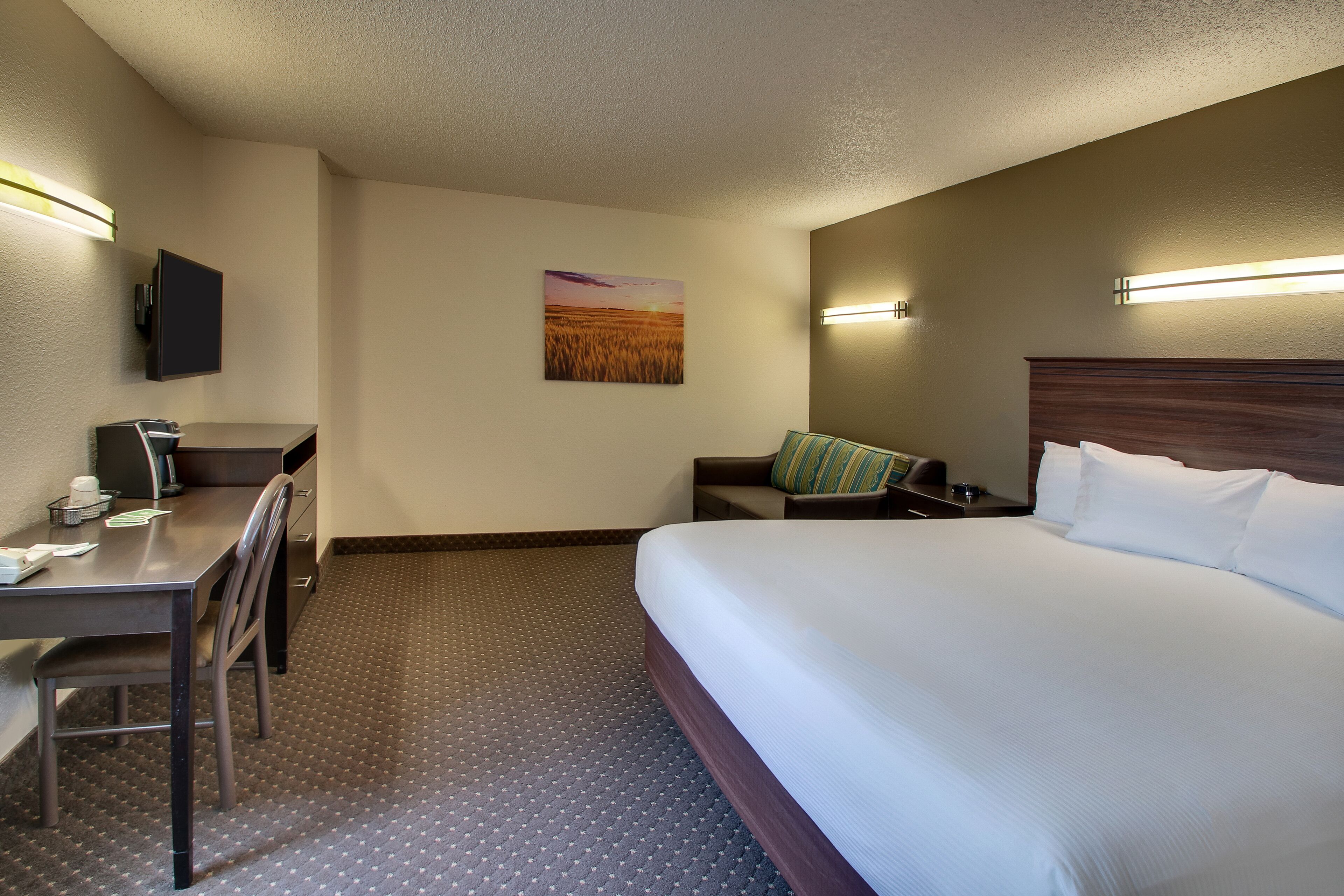 Quarto de hotel com uma cama, uma escrivaninha com cadeira, televisão na parede e um quadro de um pôr do sol.