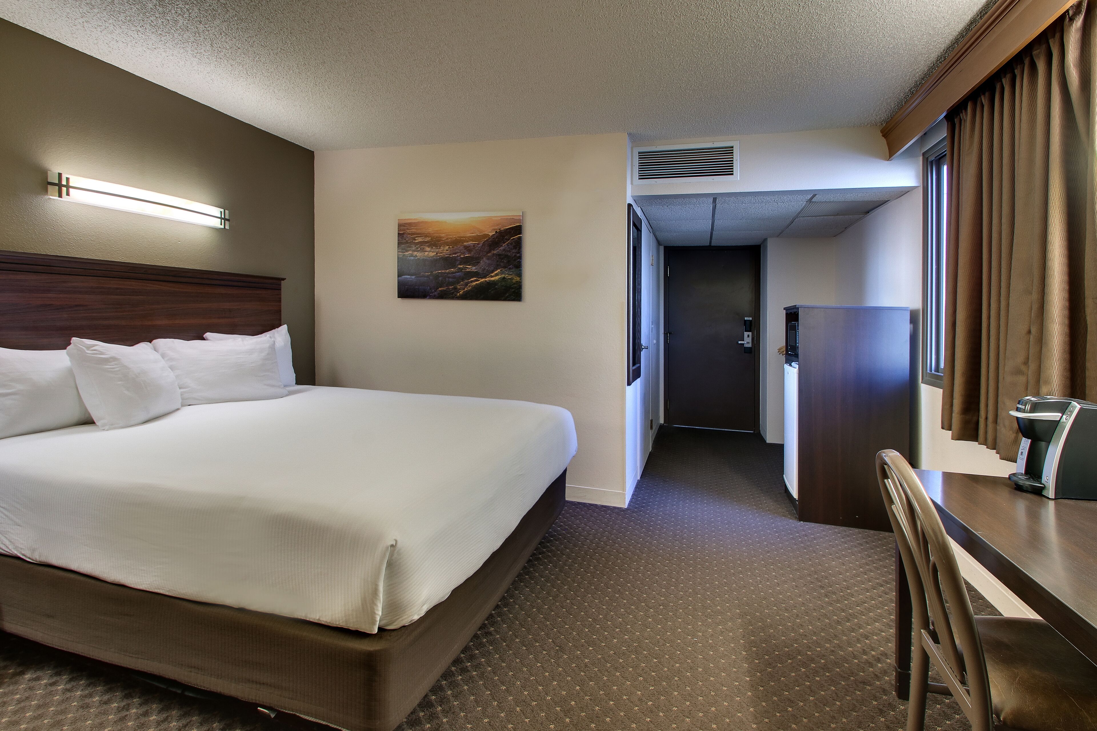 A hotel room with a large bed, a desk with a coffee maker, a painting on the wall, and a door leading to a hallway.