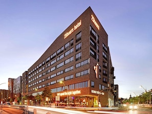 Un hotel moderno con un letrero que indica servicio 25 horas, un bar y el lema 'Pasemos la noche juntos'.