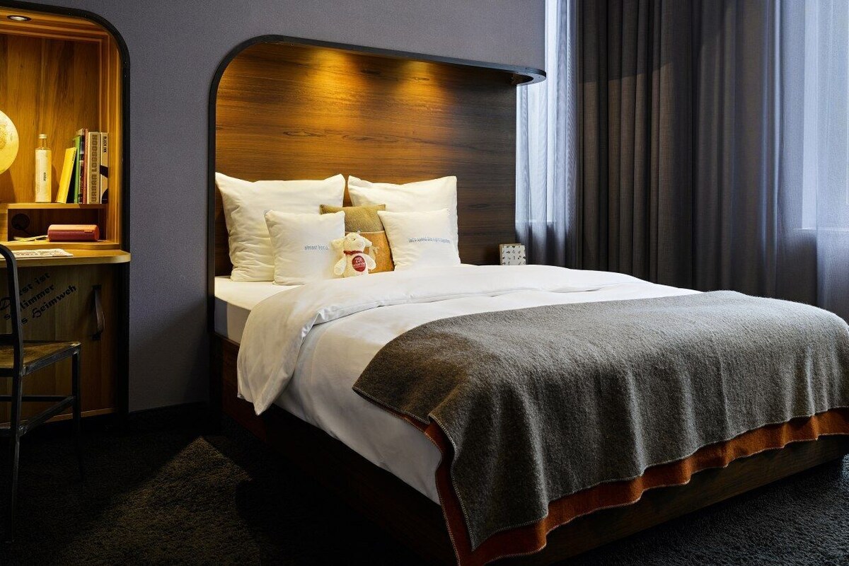 Habitación de hotel con cabecera de madera, un globo terráqueo, libros y una cama con sábanas blancas y una manta gris.