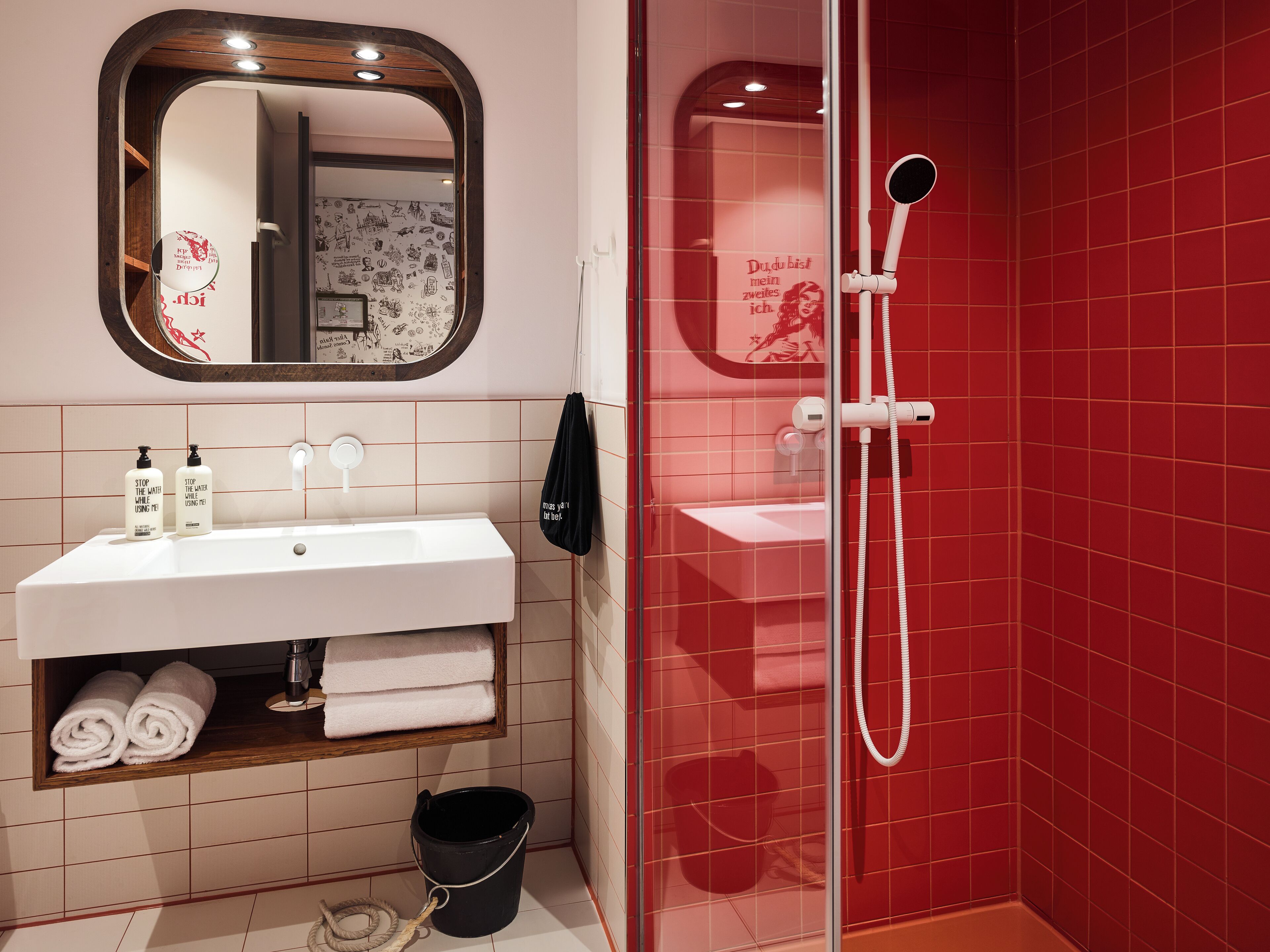 Un baño con un lavabo blanco, un espejo, una ducha con pared de azulejos rojos y un cubo negro.
