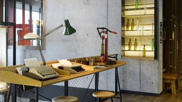 Ein Arbeitsplatz mit Schreibtisch, zwei Hocker, einer Schreibmaschine, einer Lampe und einem Weinkühler mit Flaschen.