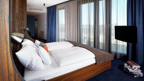 Habitación de hotel con una cama grande, un televisor de pantalla plana y vistas de la ciudad a través de amplios ventanales.