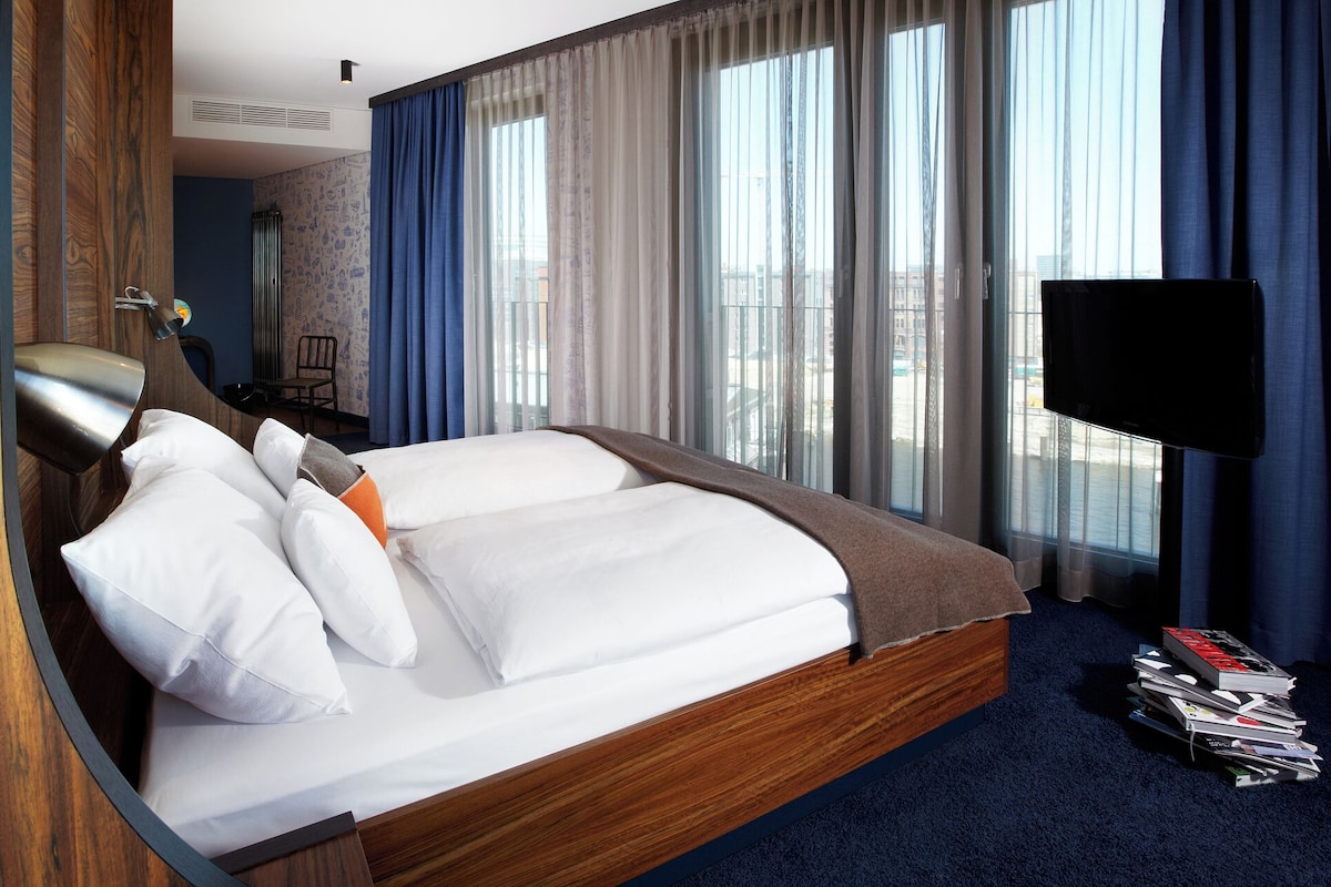 Habitación de hotel con una cama grande, un televisor de pantalla plana y vistas de la ciudad a través de amplios ventanales.