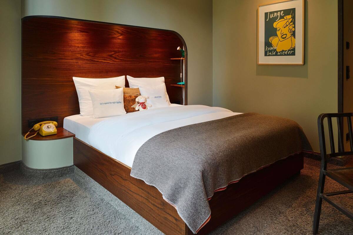 A neatly made bed with a wooden headboard, white pillows, and a grey throw blanket. A vintage telephone is on a bedside table, and a framed poster is on the wall.