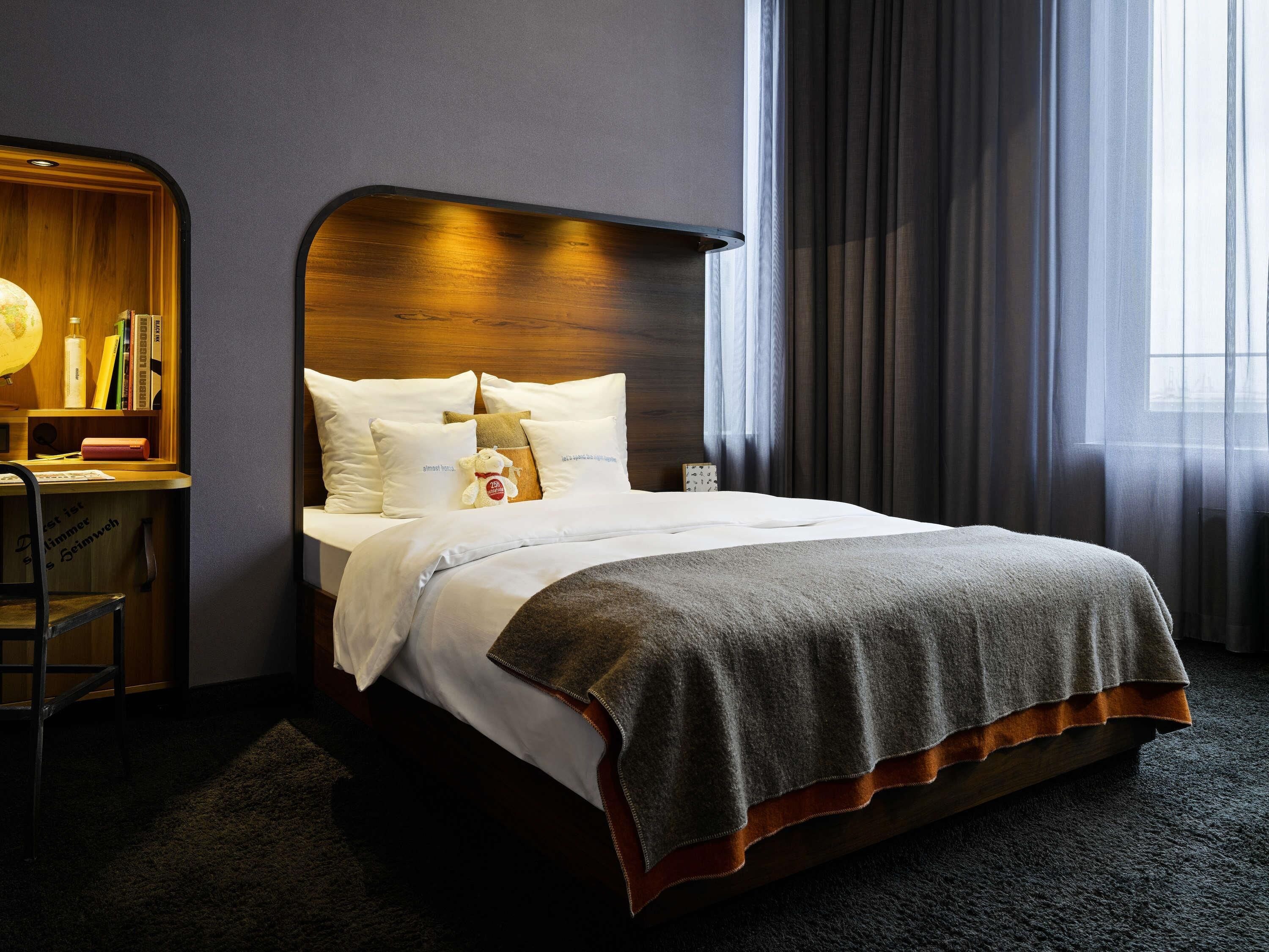 Uma cama bem arrumada com almofadas e um cobertor, uma escrivaninha de madeira com um globo e livros, e uma janela grande com cortinas.