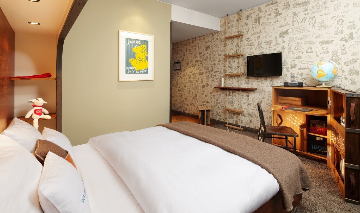 Habitación de hotel con cama, escritorio con un globo terráqueo y libros, televisor en la pared y un cartel en la pared.