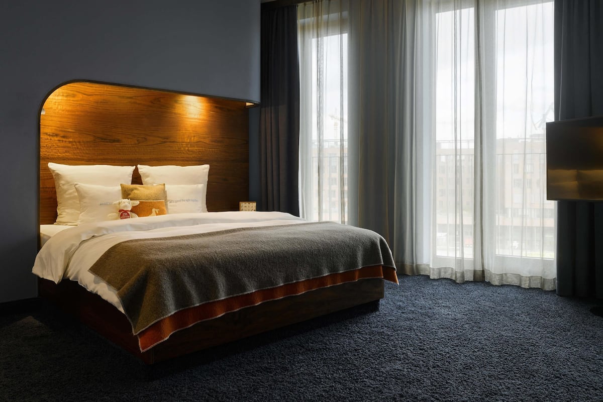 A hotel room with a large bed, a wooden headboard, a nightstand, and a view of the city through sheer curtains.