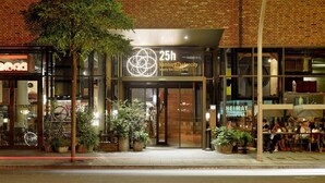 Entrada de un hotel con un letrero de 25h, puertas de vidrio y plantas en macetas.