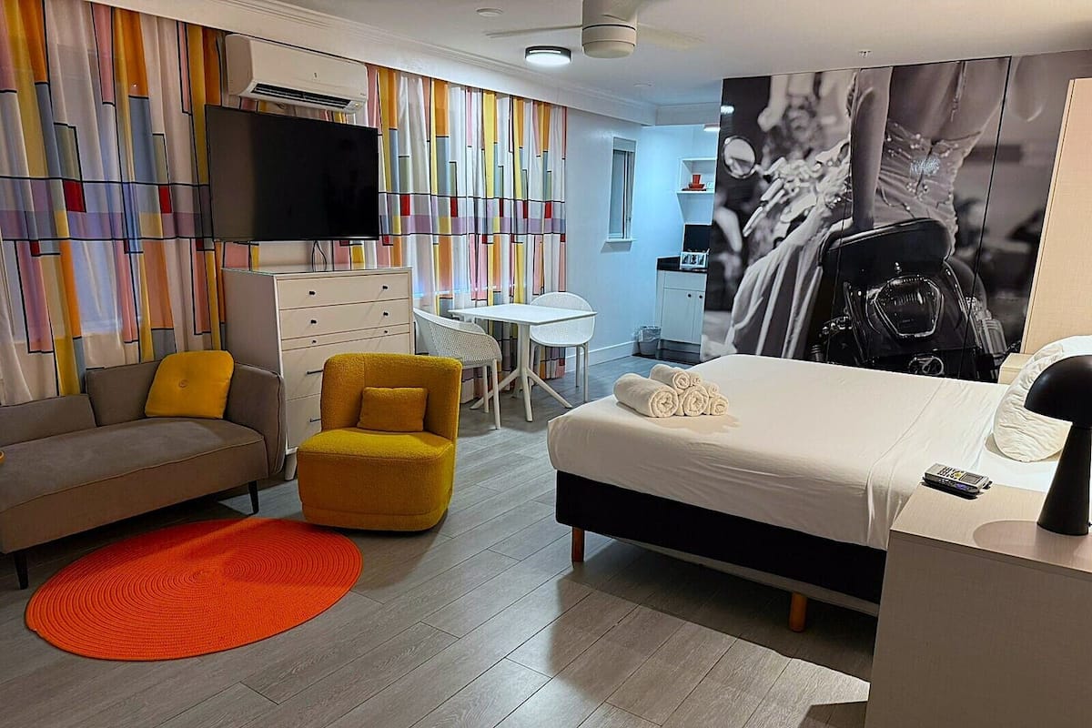 A modern hotel room with a bed, a sofa, a yellow armchair, a small table, and a TV.