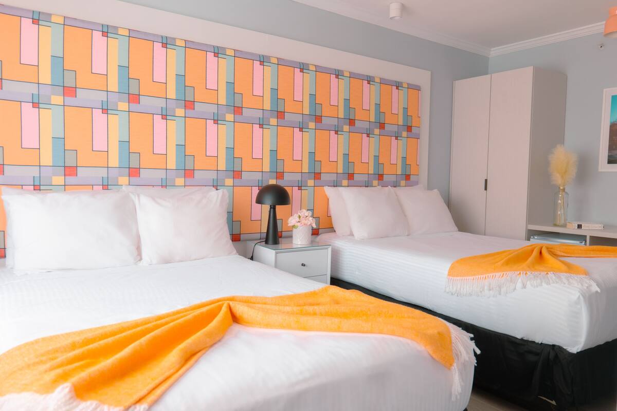 A hotel room with two beds, a colorful geometric wall art, a white wardrobe, and a nightstand with a lamp and a vase.